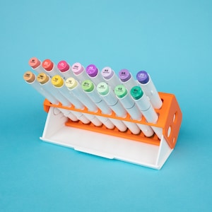 Könnte beinhalten: Ein weißer und orangefarbener Markierstift-Halter mit 16 bunten Markierstiften in einer Reihe. Die Stifte sind mit Zahlen und Buchstaben beschriftet.