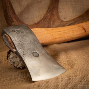 Peut inclure: Une tête de hache en métal avec un manche en bois clair. La tête de hache présente le numéro 203 estampillé et un petit emblème circulaire. La surface métallique a un aspect texturé et patiné, et le manche est en bois clair lisse.