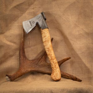 Peut inclure: Une hachette avec une tête en acier poli portant des marquages estampés et un manche en bois clair incurvé au grain distinct. La hachette est positionnée en appui contre un bois de cerf brun à plusieurs pointes.