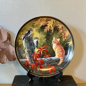 May include: A decorative ceramic plate with a fall scene featuring a kitten, two birds, and red leaves. The plate is on a black book with gold lettering.