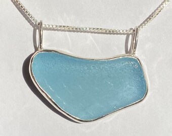 May include: A silver chain necklace with a light blue sea glass pendant. The pendant is shaped like a teardrop and has a smooth, polished surface.