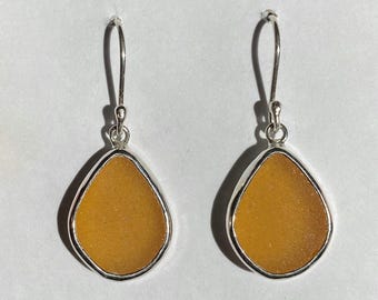 May include: A pair of dangle earrings featuring a teardrop-shaped piece of amber sea glass set in a silver frame.
