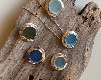 May include: Five round pendants featuring various shades of blue and green glass encased in silver settings. The pendants are displayed on a piece of driftwood.