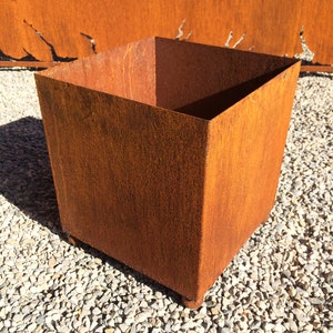Könnte beinhalten: Ein quadratischer, rostiger Metall-Pflanzkasten mit einer rauen, strukturierten Oberfläche. Der Pflanzkasten steht auf einem Bett aus kleinen, grauen Kieselsteinen.