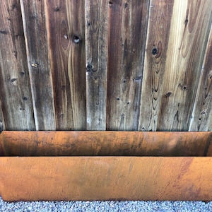 Op de afbeelding: Een rechthoekige, roestige metalen plantenbak met een licht afgeschuinde boven- en onderkant. De bak staat op een bed van kleine, grijze grindstenen.