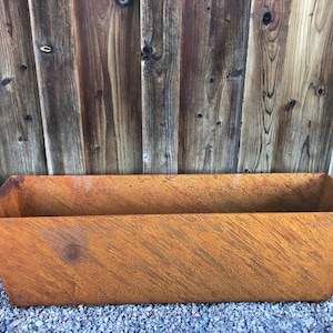 Könnte beinhalten: Ein rechteckiger, rostiger Metall-Pflanzkasten mit leicht abgeschrägter Ober- und Unterseite. Der Pflanzkasten steht auf einem Bett aus kleinen grauen Steinen.