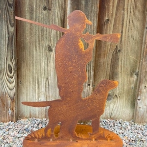 Könnte beinhalten: Eine rostige Metallsilhouette eines Jägers mit Gewehr und Hund, die auf einem runden Sockel steht. Die Silhouette ist dunkelbraun mit einer rustikalen Oberfläche.