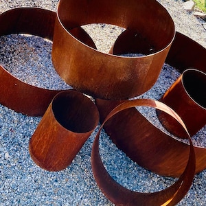Könnte beinhalten: Eine Ansammlung verwitterter, rostiger Metallringe und -zylinder in verschiedenen Größen. Die Teile sind auf einem Bett aus kleinem, grauem Kies angeordnet, wobei das natürliche Licht die strukturierten Oberflächen und warmen, erdigen Töne des Metalls hervorhebt.