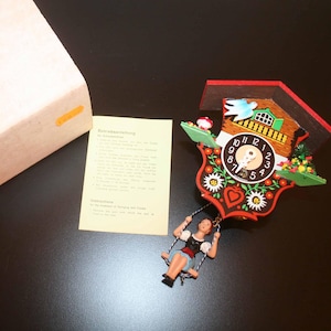 May include: A colorful cuckoo clock with a woman on a swing, a bird, and floral designs. The clock has a red roof, a black clock face, and a brown house. A box and instructions are also present.