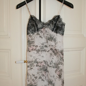 May include: A slip dress with a black lace neckline and thin straps hangs on a black hanger. The dress features a tie-dye pattern in shades of gray and pink on a white background. The dress is made of a lightweight fabric.