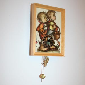 May include: A framed wall clock featuring a vintage illustration of two children. The clock has a light wood frame, a round clock face, and decorative hanging weights. The illustration depicts children in rustic clothing, carrying a basket.