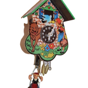 Puede incluir: Un reloj de cuco decorativo con una colorida ilustración de Caperucita Roja y el lobo. El reloj tiene un techo verde, una esfera con números negros y un péndulo con una pequeña figura oscilante. El reloj es de madera.