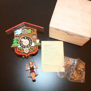 May include: A miniature cuckoo clock with a red roof, wooden facade, and a swinging figure. The clock face has black numbers and hands. Includes a cardboard box, instructions, and a small plastic bag with accessories.