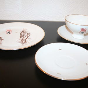 Puede incluir: Un juego de té de porcelana blanca con ribete dorado. El juego incluye una taza de té, un platillo y dos platos. Los platos y la taza de té presentan una ilustración marrón de una persona y un escudo de armas. El juego está sobre una superficie negra.