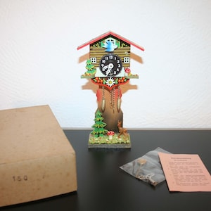 May include: A miniature cuckoo clock with a wooden facade, featuring a red roof, a black clock face, and decorative elements like a bird, trees, and a deer. The clock is accompanied by a cardboard box and a small packet with instructions.