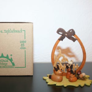 May include: Wooden Easter decoration featuring a brown bow, an oval frame, and a scene with bunnies, a duck, and eggs. The base is a yellow leaf shape. A cardboard box with green text is on the left.