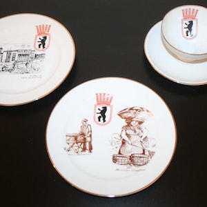 Puede incluir: Un juego de vajilla de cerámica blanca con ilustraciones de monumentos de Berlín y el escudo de armas de la ciudad. El juego incluye dos platos, una taza de té y un platillo, todos con bordes dorados. Las ilustraciones son en negro y marrón.