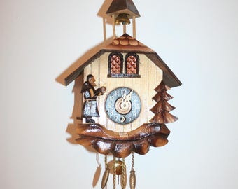 Könnte beinhalten: Eine Holzkuckucksuhr mit hellem Zifferblatt und dunkelbraunem Dach. Die Uhr hat eine kleine Glocke, eine geschnitzte Figur und einen dekorativen Baum. Das Pendel und die Gewichte der Uhr hängen unterhalb des Zifferblatts.