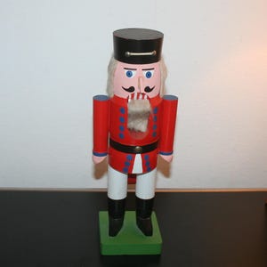 Könnte beinhalten: Eine hölzerne Nussknacker-Figur mit schwarzem Hut, roter Jacke mit blauen Knöpfen und weißer Hose. Die Figur hat ein bemaltes Gesicht mit blauen Augen, einem schwarzen Schnurrbart und einem weißen Kunstfellbart. Sie steht auf einem grünen Sockel.
