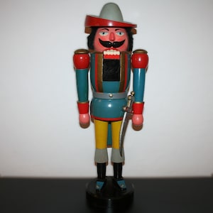 Könnte beinhalten: Eine Holz-Nussknacker-Figur in Soldatenuniform. Die Figur hat einen roten und grauen Hut, eine türkisfarbene Jacke, gelbe Hosen und schwarze Stiefel. Sie hat einen großen Schnurrbart und ein Schwert an der Seite. Der Nussknacker ist etwa 30 cm groß.