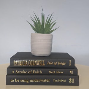 May include: A stack of three black hardcover books with gold lettering on a white surface. The top book is titled "Isle of Dogs" by Patricia Cornwell. The middle book is titled "A Stroke of Faith" by Mark Moore. The bottom book is titled "to be sung underwater" by Tom McNeal. A small potted succulent plant sits on top of the books.