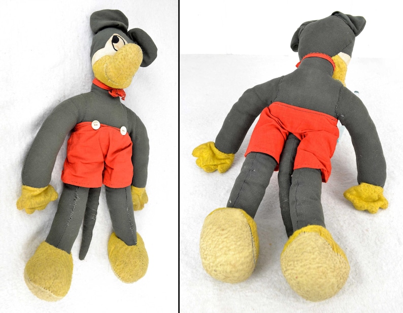 May include: A vintage plush toy of a cartoon character wearing red shorts and a black and white bow tie. The toy has a long, thin tail and is made of felt. The toy is in good condition for its age.
