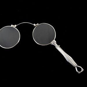 Könnte beinhalten: Antike, silberfarbene Brille mit zwei kreisförmigen Gläsern, die durch einen dünnen Draht verbunden sind. Der Griff ist aufwendig mit einem gedrehten Muster und einer Schlaufe versehen. Die Gläser sind dunkel, vor schwarzem Hintergrund.