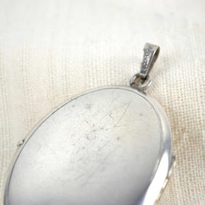 May include: Close-up of a silver oval locket with a small loop for a chain. The locket has a shiny, reflective surface with some scratches. The background is a textured, off-white fabric.