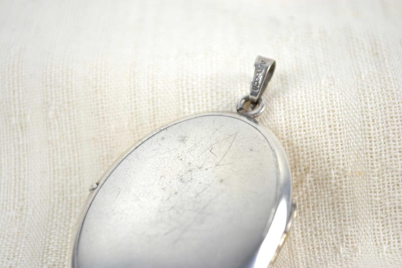 May include: Close-up of a silver oval locket with a small loop for a chain. The locket has a shiny, reflective surface with some scratches. The background is a textured, off-white fabric.