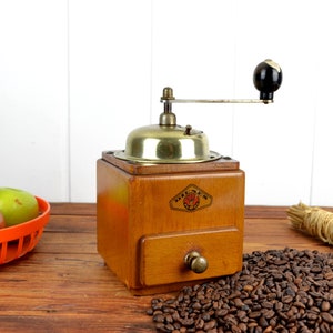 K&ouml;nnte beinhalten: Eine antike Kaffeem&uuml;hle aus Holz mit Messingdeckel und Schublade. Die M&uuml;hle hat einen schwarzen Griff und steht auf einem Holztisch mit einem Haufen Kaffeebohnen davor.