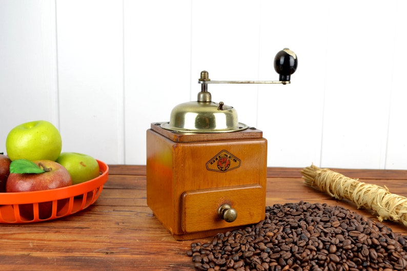 K&ouml;nnte beinhalten: Eine antike Kaffeem&uuml;hle aus Holz mit Messingdeckel und Schublade. Die M&uuml;hle hat einen schwarzen Griff und steht auf einem Holztisch mit einem Haufen Kaffeebohnen davor.