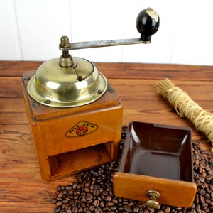 K&ouml;nnte beinhalten: Eine antike Kaffeem&uuml;hle aus Holz mit Messingdeckel und einer Schublade zum Auffangen des Kaffeesatzes. Die M&uuml;hle hat einen Kurbelgriff und ein Etikett mit der Aufschrift "Dresden". Die M&uuml;hle steht auf einer Holzoberfl&auml;che mit einem Haufen Kaffeebohnen davor.