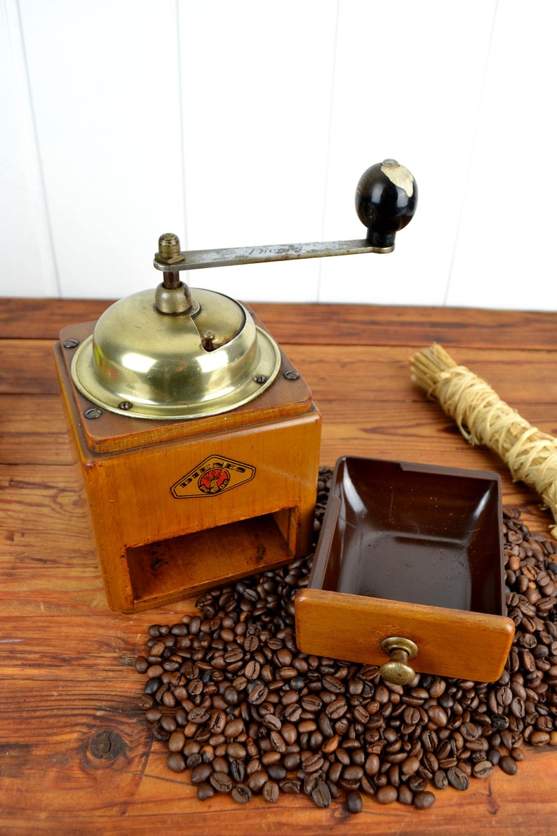 K&ouml;nnte beinhalten: Eine antike Kaffeem&uuml;hle aus Holz mit Messingdeckel und einer Schublade zum Auffangen des Kaffeesatzes. Die M&uuml;hle hat einen Kurbelgriff und ein Etikett mit der Aufschrift "Dresden". Die M&uuml;hle steht auf einer Holzoberfl&auml;che mit einem Haufen Kaffeebohnen davor.