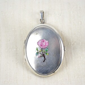 May include: Oval silver locket with a pink rose design. The locket has a hinged opening and a small loop at the top for a chain. The rose has green leaves and a brown stem. The locket is displayed on a white textured surface.