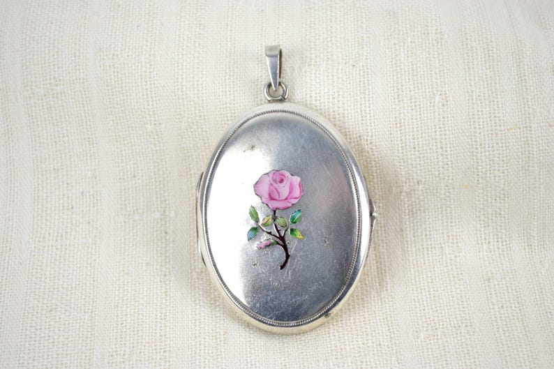 May include: Oval silver locket with a pink rose design. The locket has a hinged opening and a small loop at the top for a chain. The rose has green leaves and a brown stem. The locket is displayed on a white textured surface.