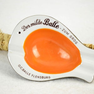Puede incluir: Cenicero de cerámica blanca con interior naranja. El cenicero tiene forma de pez y tiene el texto "Der milde Balle - zum Grog O.C. Balle Flensburg" impreso en la parte inferior.