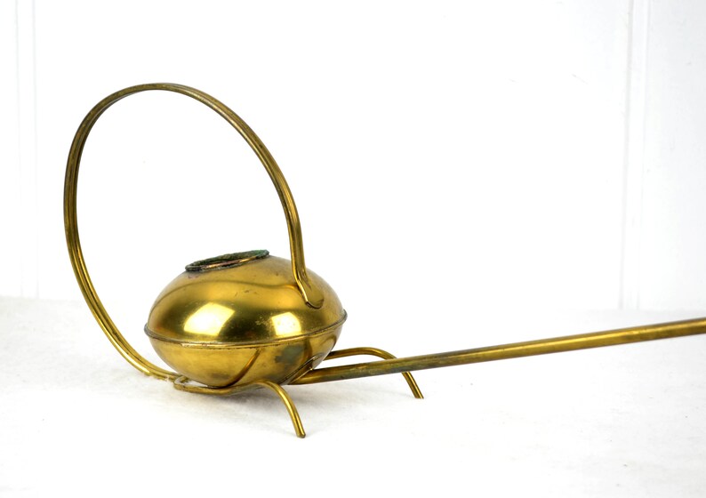 May include: A brass watering can with a long spout and a curved handle. The watering can has a round body and a decorative base.
