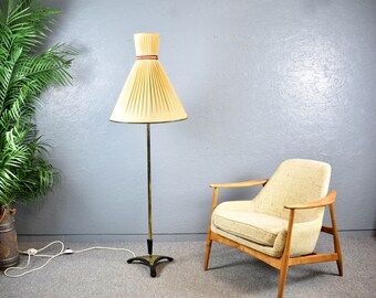 Könnte beinhalten: Eine Stehlampe aus Messing mit einem gefalteten weißen Lampenschirm und einem braunen Besatz. Die Lampe hat einen dreibeinigen Sockel und steht neben einem beigen Sessel.