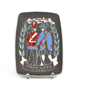 May include: A black ceramic plate featuring a colourful illustration of two soldiers standing in front of a fortress. The plate has a white banner with the text "FREDRIKSTEN NORGE".