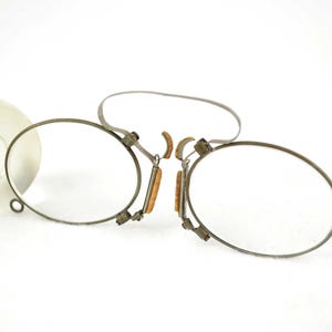 Könnte beinhalten: Antike Brille mit runden Gläsern und einem silberfarbenen Metallrahmen. Die Brille hat kleine, rechteckige Nasenpads und ein dünnes, flexibles Band. Eine transparente Glasflasche ist im Hintergrund zu sehen.