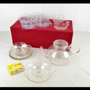 May include: A clear glass tea set comprising a teapot, cups, and saucers. The set includes a teapot with a handle and lid, several cups with handles, and matching saucers. The set is displayed on a white surface with a red box in the background.