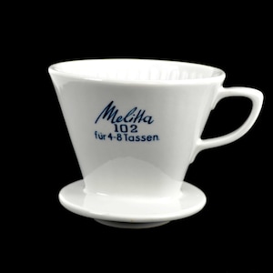 May include: White ceramic Melitta 102 pour-over coffee maker with a handle and a small base. The words "Melitta 102 für 4-8 Tassen" are printed in blue on the front. The coffee maker is designed for brewing 4-8 cups of coffee.
