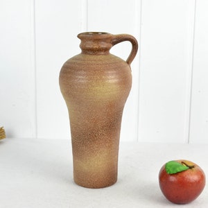 May include: A brown ceramic pitcher with a textured surface and a handle. The pitcher is on a white surface with a red apple.