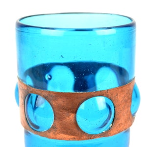 May include: A turquoise blue glass drinking glass with a copper band around the middle. The band has three circular cutouts that reveal the glass underneath.