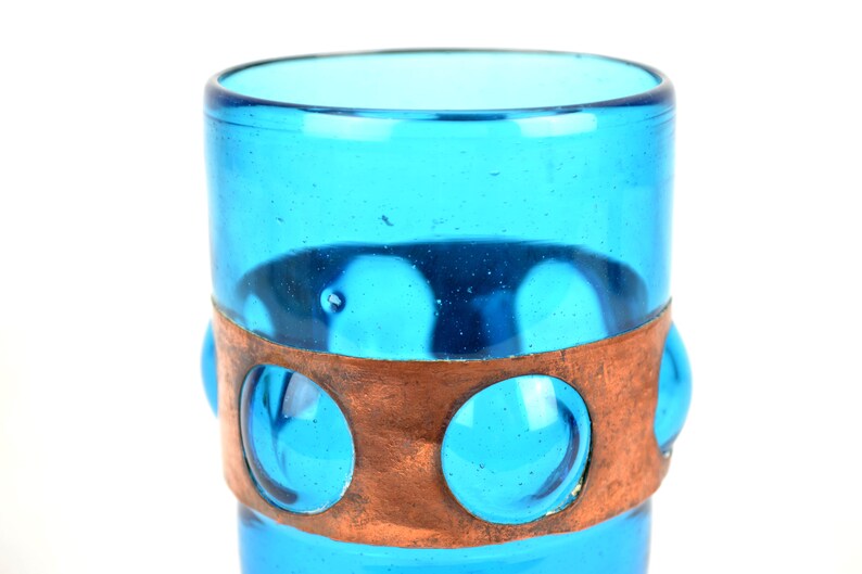 May include: A turquoise blue glass drinking glass with a copper band around the middle. The band has three circular cutouts that reveal the glass underneath.