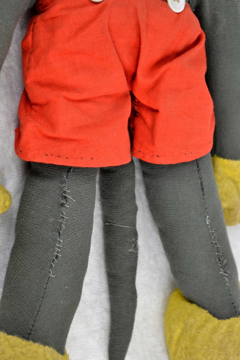 May include: A vintage plush toy of Mickey Mouse. The toy is wearing red shorts and has a black tail. The toy is made of grey fabric and has yellow shoes.