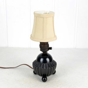 May include: A small, black table lamp with a cream lampshade. The lamp has a round base with a ribbed design and three small feet. A brown cable extends from the base.