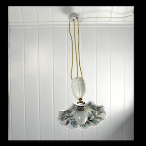 Puede incluir: Una lámpara colgante de cerámica blanca con un diseño floral. La lámpara tiene una pantalla blanca con detalles florales azules y verdes. La lámpara está colgada de un techo blanco.