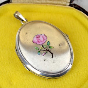 May include: Oval silver locket with a pink rose design on the front. The locket is set on a yellow velvet surface inside a black and brown case. The rose has green leaves and a black stem.