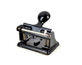May include: A black metal hole punch with a lever handle and a spring-loaded mechanism. The punch is designed to create holes in paper.